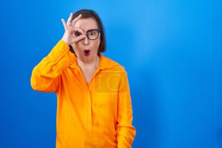 Téléchargez les photos : Femme hispanique du Moyen Âge portant des lunettes debout sur fond bleu faisant un geste correct choqué par le visage surpris, les yeux regardant à travers les doigts. expression incrédule. - en image libre de droit