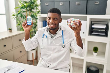 Foto de Hombre médico afroamericano usando medidor de glucosa sonriendo mirando hacia un lado y mirando hacia otro pensando. - Imagen libre de derechos