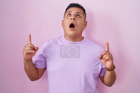 Téléchargez les photos : Young hispanic man standing over pink background amazed and surprised looking up and pointing with fingers and raised arms. - en image libre de droit