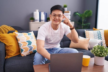 Téléchargez les photos : Jeune homme asiatique utilisant un ordinateur portable à la maison assis sur le canapé souriant pointant vers la tête avec un doigt, grande idée ou pensée, bonne mémoire - en image libre de droit