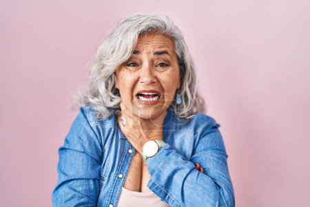 Foto de Mujer de mediana edad con el pelo gris de pie sobre el fondo rosa gritando sofocar porque estrangulamiento doloroso. problema de salud. concepto de asfixia y suicidio. - Imagen libre de derechos