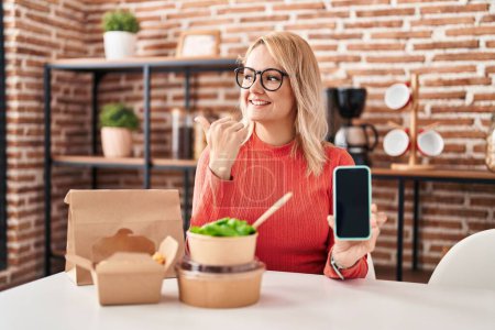 Photo for Blonde woman eating take away food showing smartphone screen pointing thumb up to the side smiling happy with open mouth - Royalty Free Image