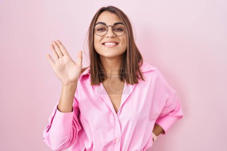 Téléchargez les photos : Jeune femme hispanique portant des lunettes debout sur fond rose renonçant à dire bonjour heureux et souriant, geste de bienvenue amical - en image libre de droit