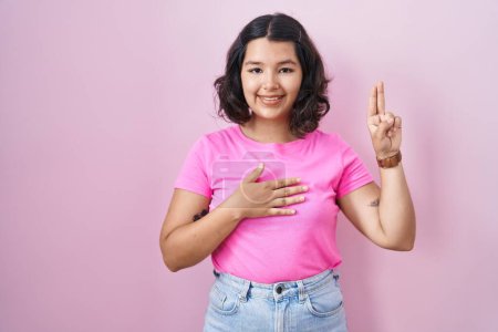 Téléchargez les photos : Jeune femme hispanique debout sur fond rose souriant jurer avec la main sur la poitrine et les doigts vers le haut, faisant un serment de fidélité promesse - en image libre de droit