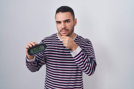Photo for Young hispanic man wearing pyjama holding alarm clock serious face thinking about question with hand on chin, thoughtful about confusing idea - Royalty Free Image