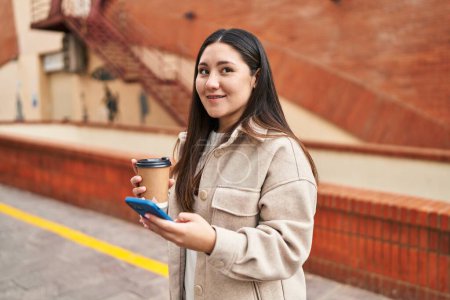 Téléchargez les photos : Young hispanic woman using smartphone drinking coffee at street - en image libre de droit