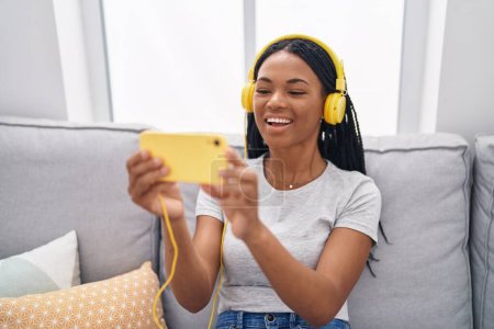 Téléchargez les photos : Femme afro-américaine regarder la vidéo sur smartphone assis sur le canapé à la maison - en image libre de droit