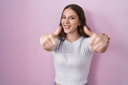 Téléchargez les photos : Jeune fille hispanique debout sur fond rose approuvant faire un geste positif avec la main, pouces levés souriant et heureux pour le succès. geste gagnant. - en image libre de droit