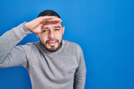 Photo for Hispanic man standing over blue background very happy and smiling looking far away with hand over head. searching concept. - Royalty Free Image