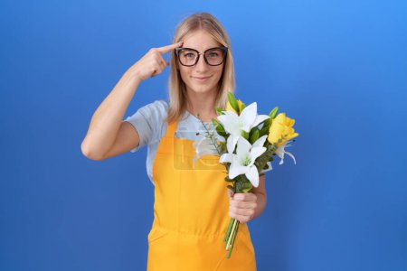 Téléchargez les photos : Jeune femme caucasienne portant tablier de fleuriste tenant des fleurs souriant pointant vers la tête avec un doigt, grande idée ou pensée, bonne mémoire - en image libre de droit