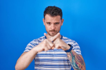 Téléchargez les photos : Young hispanic man standing over blue background rejection expression crossing fingers doing negative sign - en image libre de droit