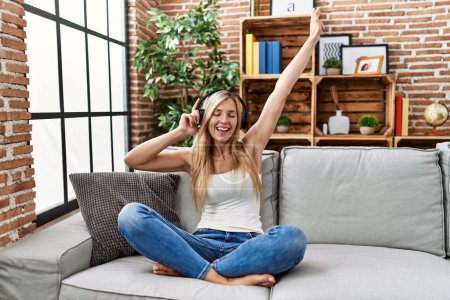 Téléchargez les photos : Jeune femme blonde écoutant de la musique et dansant assis sur le canapé à la maison - en image libre de droit