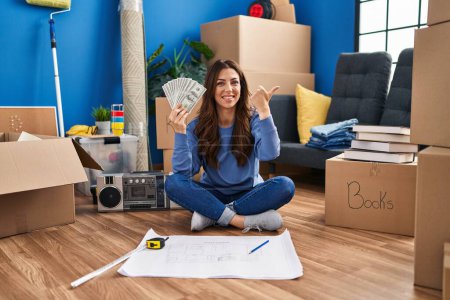 Photo for Young brunette woman sitting on the floor at new home holding money smiling happy pointing with hand and finger to the side - Royalty Free Image