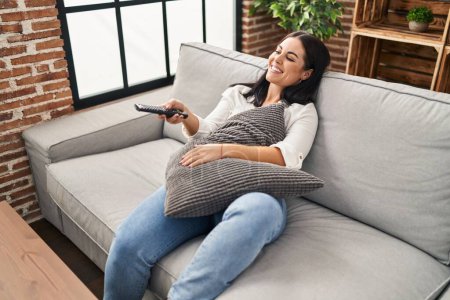 Photo for Young beautiful hispanic woman watching tv sitting on sofa at home - Royalty Free Image