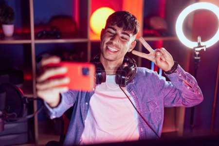 Foto de Joven hombre hispano streamer sonriendo confiado hacer selfie por teléfono inteligente en la sala de juegos - Imagen libre de derechos