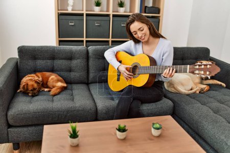 Téléchargez les photos : Jeune femme hispanique jouant de la guitare classique assise sur un canapé avec des chiens à la maison - en image libre de droit