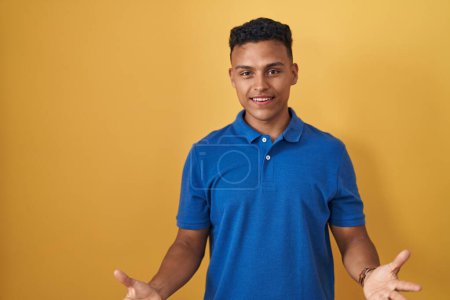 Foto de Joven hispano de pie sobre fondo amarillo sonriente alegre con los brazos abiertos como bienvenida amistosa, saludos positivos y confiados - Imagen libre de derechos