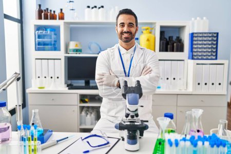 Téléchargez les photos : Jeune homme hispanique avec barbe travaillant au laboratoire scientifique heureux visage souriant avec les bras croisés regardant la caméra. personne positive. - en image libre de droit
