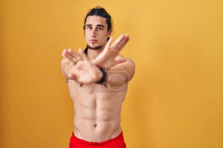 Photo for Hispanic man with long hair standing shirtless over yellow background rejection expression crossing arms and palms doing negative sign, angry face - Royalty Free Image