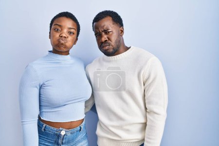 Foto de Joven pareja afroamericana de pie sobre fondo azul hinchando mejillas con cara divertida. boca hinchada de aire, expresión loca. - Imagen libre de derechos