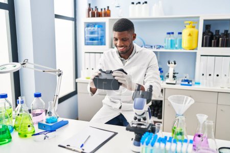 Photo for Young african american man wearing scientist uniform holding virtual reality glasses at laboratory - Royalty Free Image