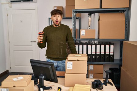 Téléchargez les photos : Arab man with beard working at small business commerce holding bar code scanner scared and amazed with open mouth for surprise, disbelief face - en image libre de droit