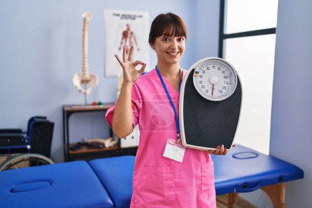 Photo for Young brunette woman as nutritionist holding weighing machine doing ok sign with fingers, smiling friendly gesturing excellent symbol - Royalty Free Image