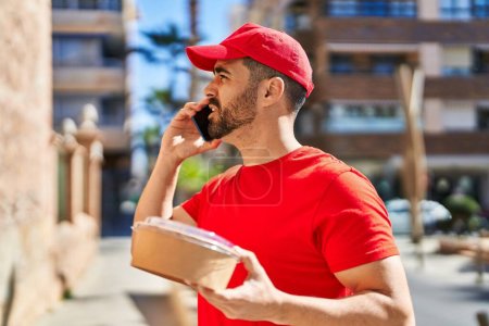 Photo for Young hispanic man courier talking on the smartphone holding package at street - Royalty Free Image