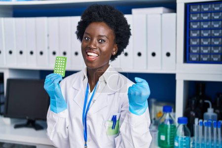 Foto de Mujer joven africana trabajando en laboratorio científico sosteniendo píldoras anticonceptivas gritando orgullosa, celebrando la victoria y el éxito muy emocionada con los brazos levantados - Imagen libre de derechos
