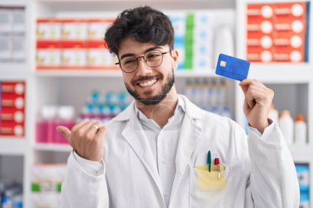 Foto de Hombre hispano con barba trabajando en farmacia sosteniendo tarjeta de crédito apuntando con el pulgar hacia un lado sonriendo feliz con la boca abierta - Imagen libre de derechos