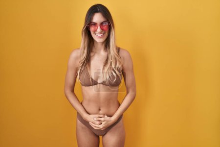 Photo for Young hispanic woman wearing bikini over yellow background with hands together and crossed fingers smiling relaxed and cheerful. success and optimistic - Royalty Free Image