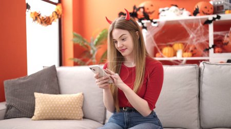 Téléchargez les photos : Jeune femme blonde souriante portant costume diable en utilisant un smartphone à la maison - en image libre de droit