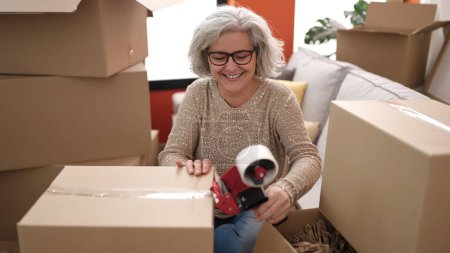 Foto de Middle age woman with grey hair smiling confident packing cardboard box at new home - Imagen libre de derechos