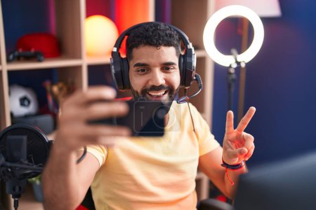 Foto de Joven árabe hombre streamer sonriendo confiado hacer selfie por teléfono inteligente en la sala de juegos - Imagen libre de derechos