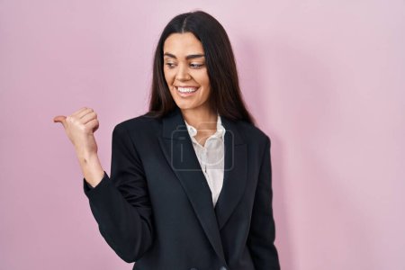 Téléchargez les photos : Jeune femme brune portant un style d'affaires sur fond rose souriant avec un visage heureux regardant et pointant vers le côté avec le pouce vers le haut. - en image libre de droit