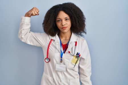 Téléchargez les photos : Jeune femme afro-américaine portant l'uniforme de médecin et stéthoscope personne forte montrant muscle du bras, confiant et fier de la puissance - en image libre de droit