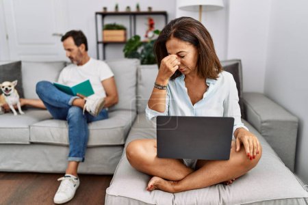 Foto de Hispanic middle age couple at home, woman using laptop tired rubbing nose and eyes feeling fatigue and headache. stress and frustration concept. - Imagen libre de derechos