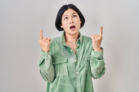 Téléchargez les photos : Young asian woman standing over white background amazed and surprised looking up and pointing with fingers and raised arms. - en image libre de droit