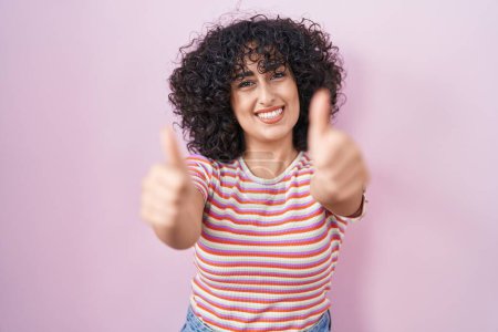 Foto de Joven mujer de Oriente Medio de pie sobre fondo rosa aprobando hacer gesto positivo con la mano, pulgares hacia arriba sonriendo y feliz por el éxito. gesto ganador. - Imagen libre de derechos
