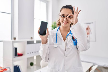 Téléchargez les photos : Jeune médecin hispanique femme tenant smartphone montrant écran souriant heureux faire ok signe avec la main sur les yeux en regardant à travers les doigts - en image libre de droit