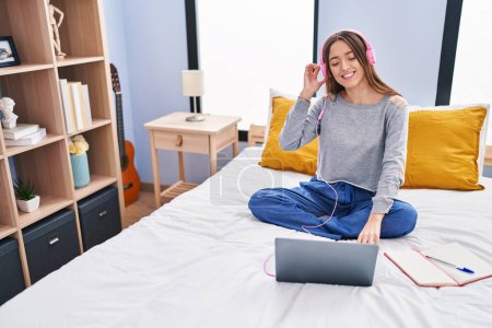 Téléchargez les photos : Jeune belle étudiante hispanique écoutant de la musique vto étudier sur le lit dans la chambre - en image libre de droit