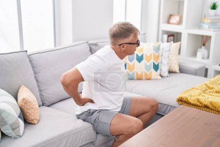 Téléchargez les photos : Homme aux cheveux gris d'âge moyen souffrant de maux de dos assis sur le canapé à la maison - en image libre de droit