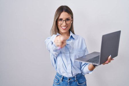 Téléchargez les photos : Jeune femme travaillant à l'aide d'un ordinateur portable pointant vers vous et la caméra avec les doigts, souriant positif et joyeux - en image libre de droit