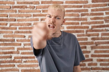 Téléchargez les photos : Jeune homme caucasien debout sur le mur de briques pointant mécontent et frustré à la caméra, en colère et furieux contre vous - en image libre de droit