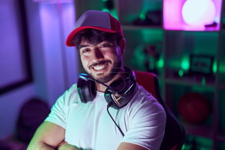 Téléchargez les photos : Jeune homme hispanique banderole souriant confiant assis avec les bras croisés geste à la salle de jeu - en image libre de droit