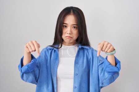 Téléchargez les photos : Jeune femme chinoise debout sur fond blanc pointant vers le bas regardant triste et contrarié, indiquant la direction avec les doigts, malheureux et déprimé. - en image libre de droit