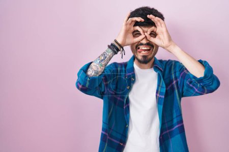Photo for Young hispanic man with beard standing over pink background doing ok gesture like binoculars sticking tongue out, eyes looking through fingers. crazy expression. - Royalty Free Image