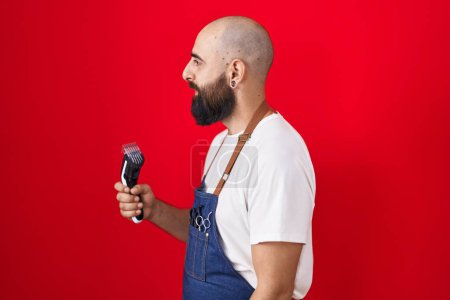 Photo for Young hispanic man with beard and tattoos wearing barber apron holding razor looking to side, relax profile pose with natural face and confident smile. - Royalty Free Image
