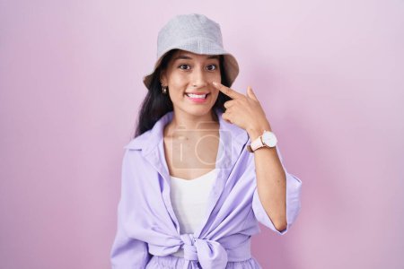 Foto de Young hispanic woman standing over pink background wearing hat pointing with hand finger to face and nose, smiling cheerful. beauty concept - Imagen libre de derechos