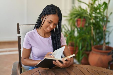 Foto de African american woman reading book sitting on table at home terrace - Imagen libre de derechos